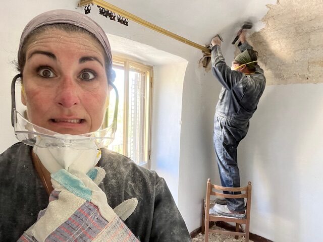 Woman covered in dust wearing protective gear during an Italian home renovation, while another person removes plaster from an old Italian ceiling using hand tools