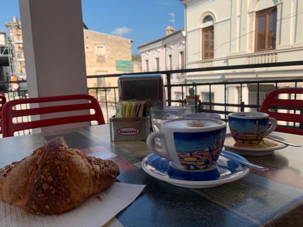 A cappuccino and cornetto on a small table in an Italian piazza, with the lively square and people passing by in the background.