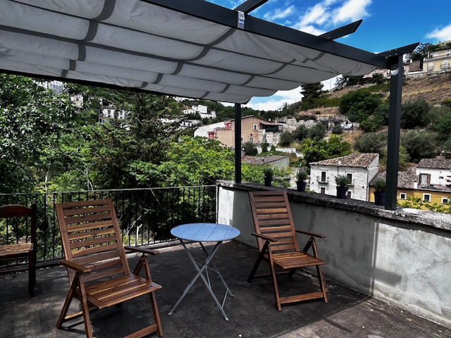 Rooftop terrace with a shaded gazebo, two wooden chairs, and a small round table, overlooking a scenic hillside village.