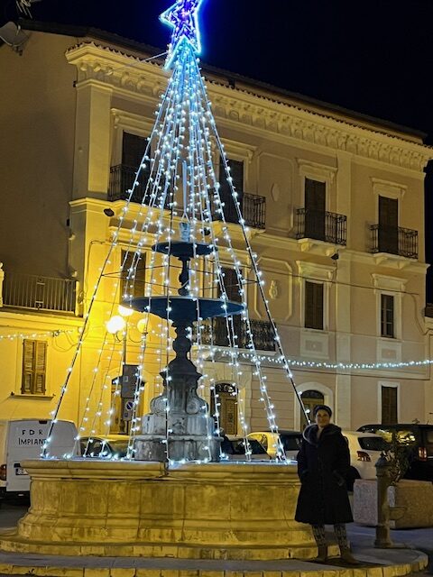 Standing warmly bundled in the piazza on a cold holiday night, surrounded by festive lights and a lively holiday crowd.