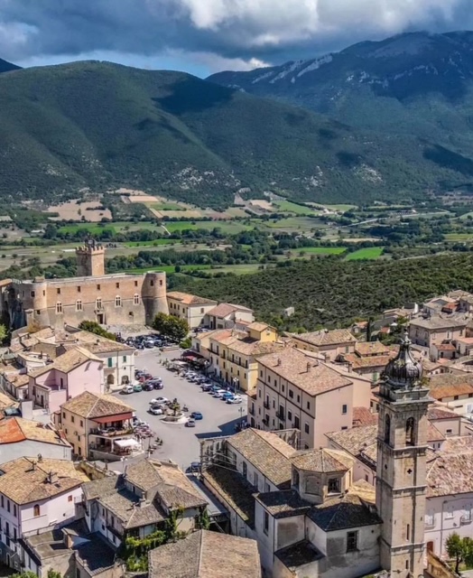 A wide view from above of the entire piazza, capturing the bustling atmosphere with people gathering around cafes, restaurants, and the central fountain amidst historic buildings.