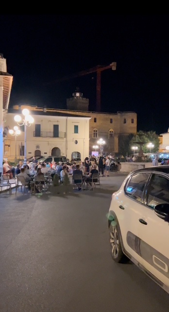 A group gathered at night in an Italian piazza, enjoying conversation and laughter under warm lights with the surrounding historic buildings softly illuminated.