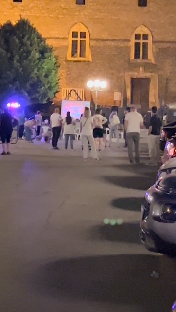 A group gathered at night in an Italian piazza, enjoying conversation and laughter under warm lights with the surrounding historic buildings softly illuminated.