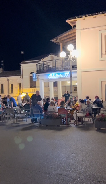 A group gathered at night in an Italian piazza, enjoying conversation and laughter under warm lights with the surrounding historic buildings softly illuminated.