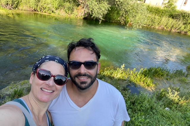  A clear, emerald-green Tirino River flows through a verdant valley lined with ancient willows and poplars under a bright blue sky, with sunlight illuminating the transparent water.