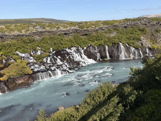 Icelandic waterfall flowing into a river surrounded by rugged green landscape.